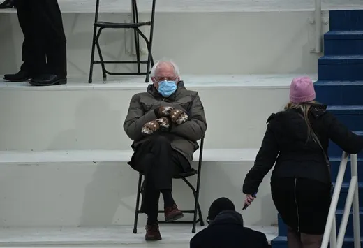 Bernie Sanders sitting on a folding chair with legs crossed, in a parka and patterned mittens