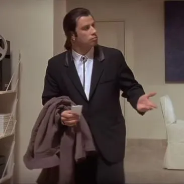 John Travolta in a black suit standing in a room, looking around in confusion