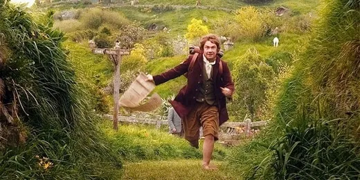 Bilbo Baggins running joyfully across a green Shire field, holding a contract