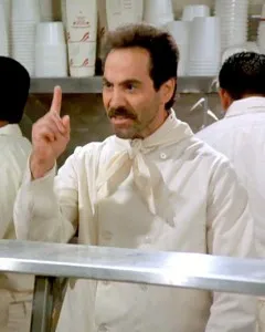 A stern man in a white chef's uniform pointing behind a soup counter