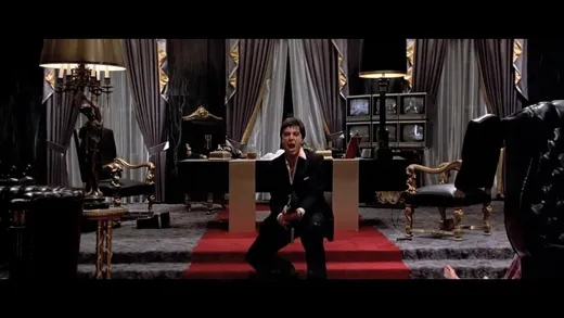 Tony Montana in a white suit at the top of a grand staircase, holding a large gun
