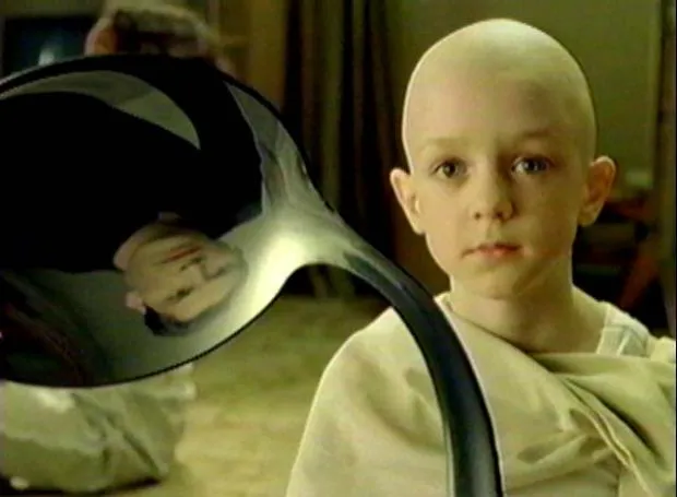 A bald boy in robes holding a bent spoon, reflected in the bowl