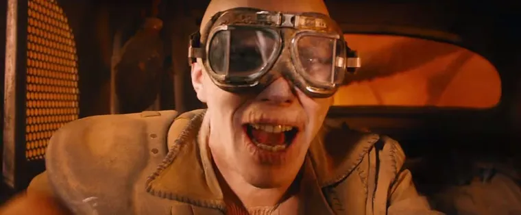 Nux, a pale war boy with goggles, grinning wildly in a car